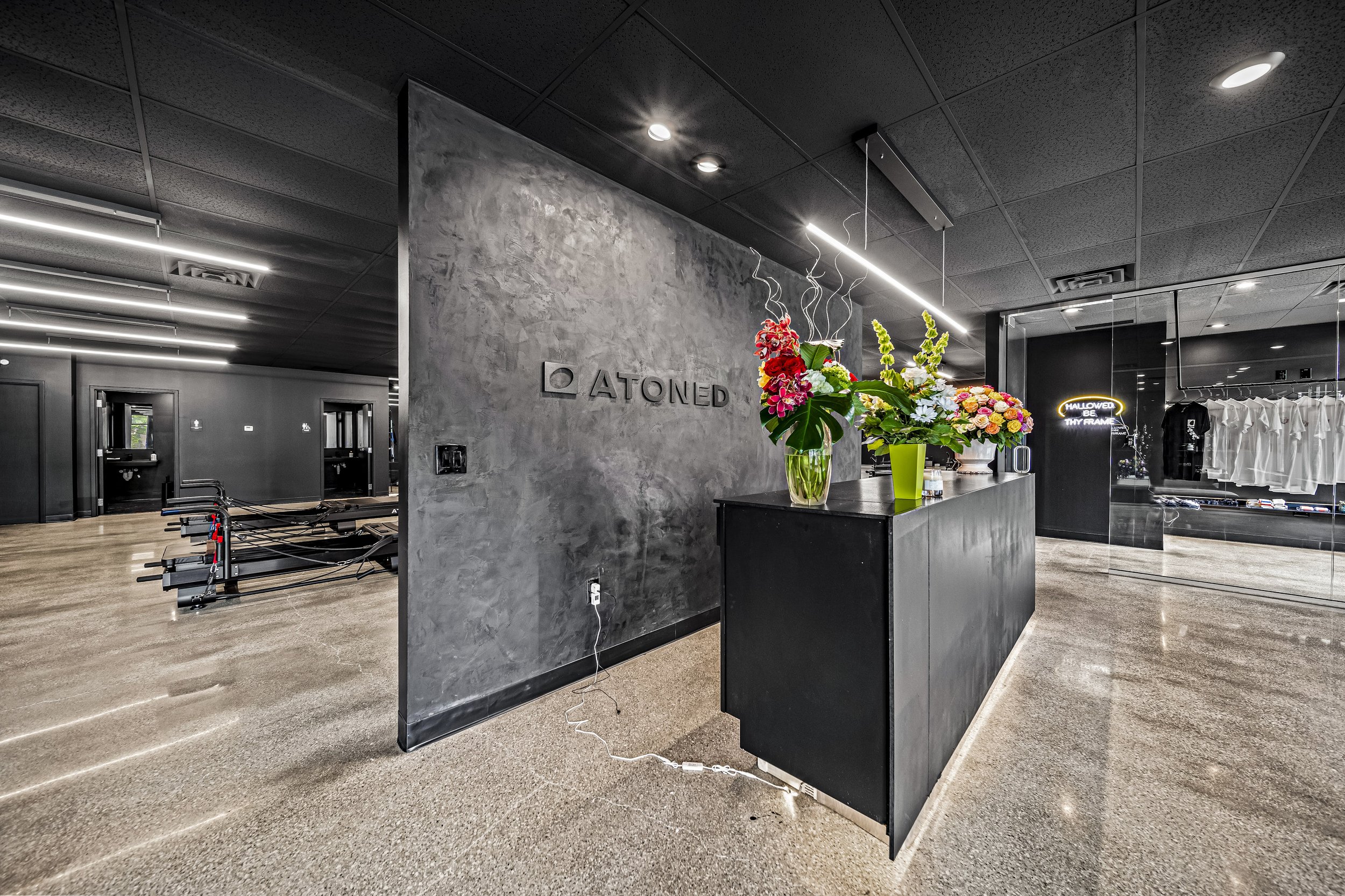 Wide-angle reception area with concrete branded feature wall, floral arrangements, and views through to reformer studio and retail — commercial fitness build-out by Skoda Construction, Northeast Ohio