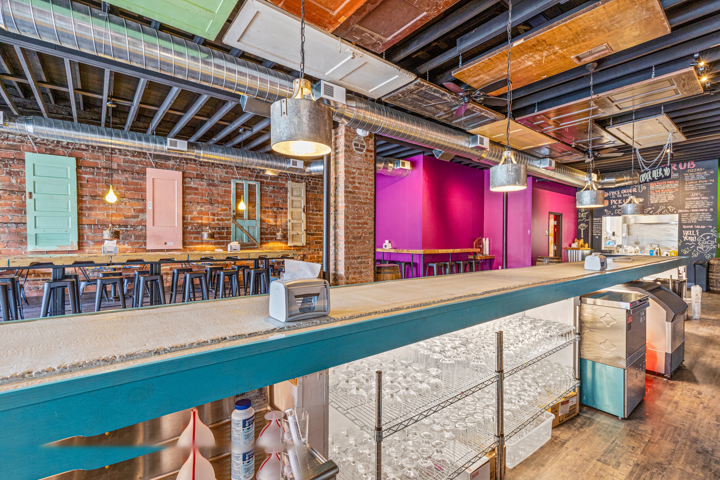 Elevated view of brewery interior with teal bar, colorful vintage doors on brick walls, and industrial pendant lighting — VooDoo Brewery by Skoda Construction