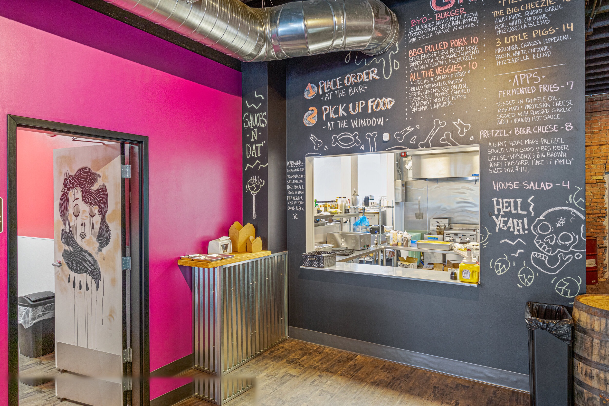 Food order window with chalkboard menu, magenta accent wall, voodoo doll door art, and corrugated metal counter — VooDoo Brewery by Skoda Construction