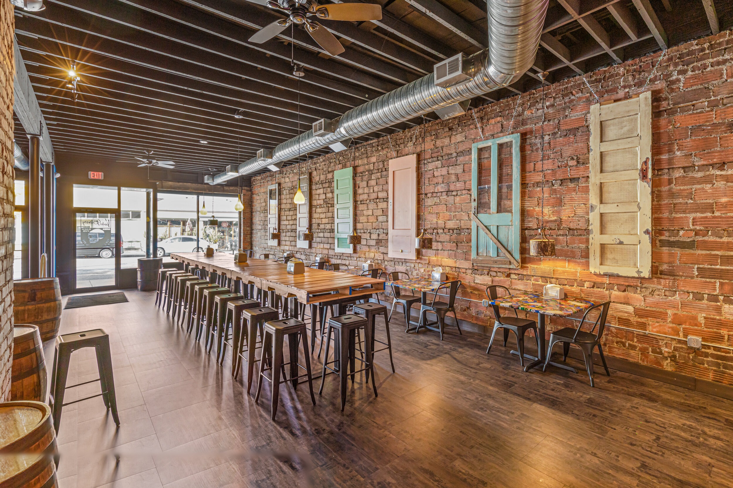 Industrial-rustic dining hall with communal wood tables, metal stools, exposed brick wall, and reclaimed shutters — VooDoo Brewery by Skoda Construction