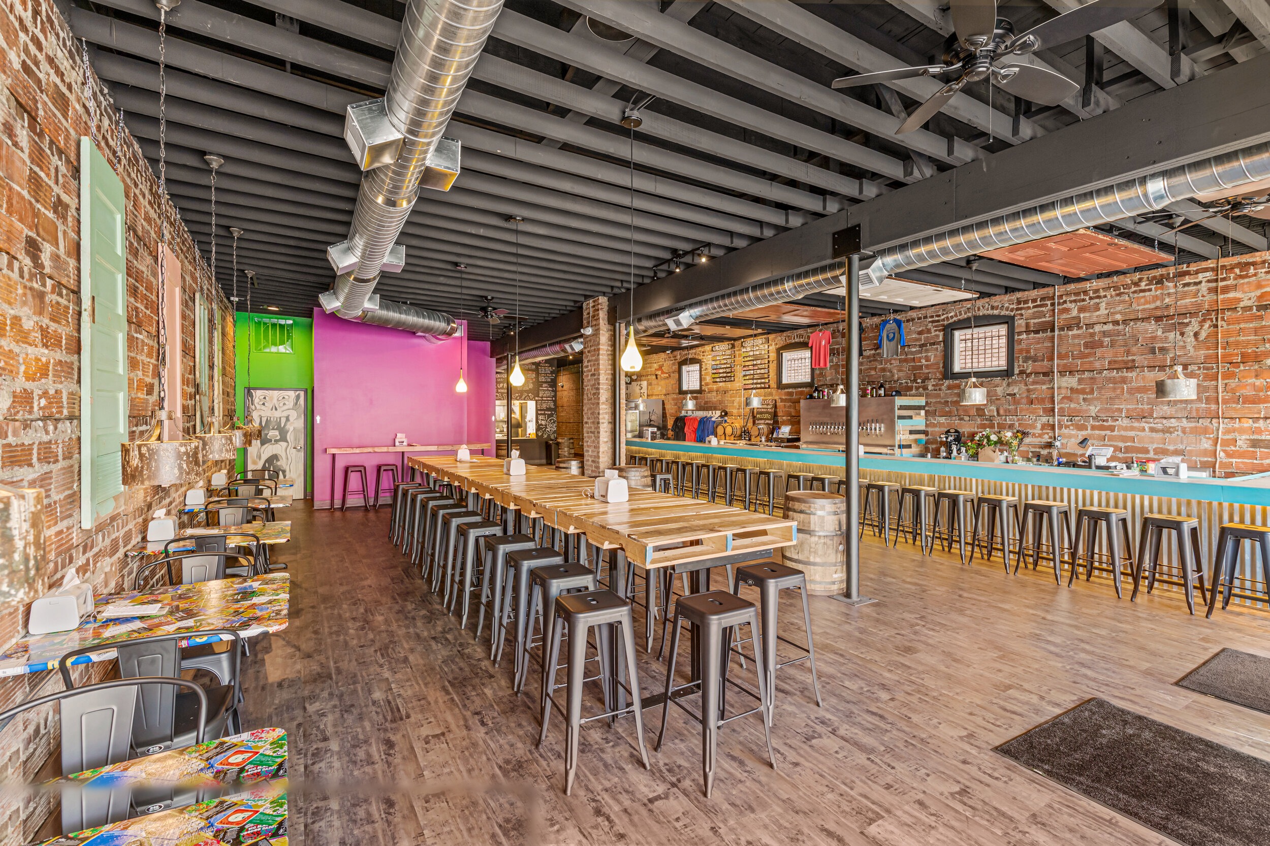 Full brewery interior with communal tables, magenta accent wall, teal bar, and exposed ductwork under dark ceiling — VooDoo Brewery by Skoda Construction
