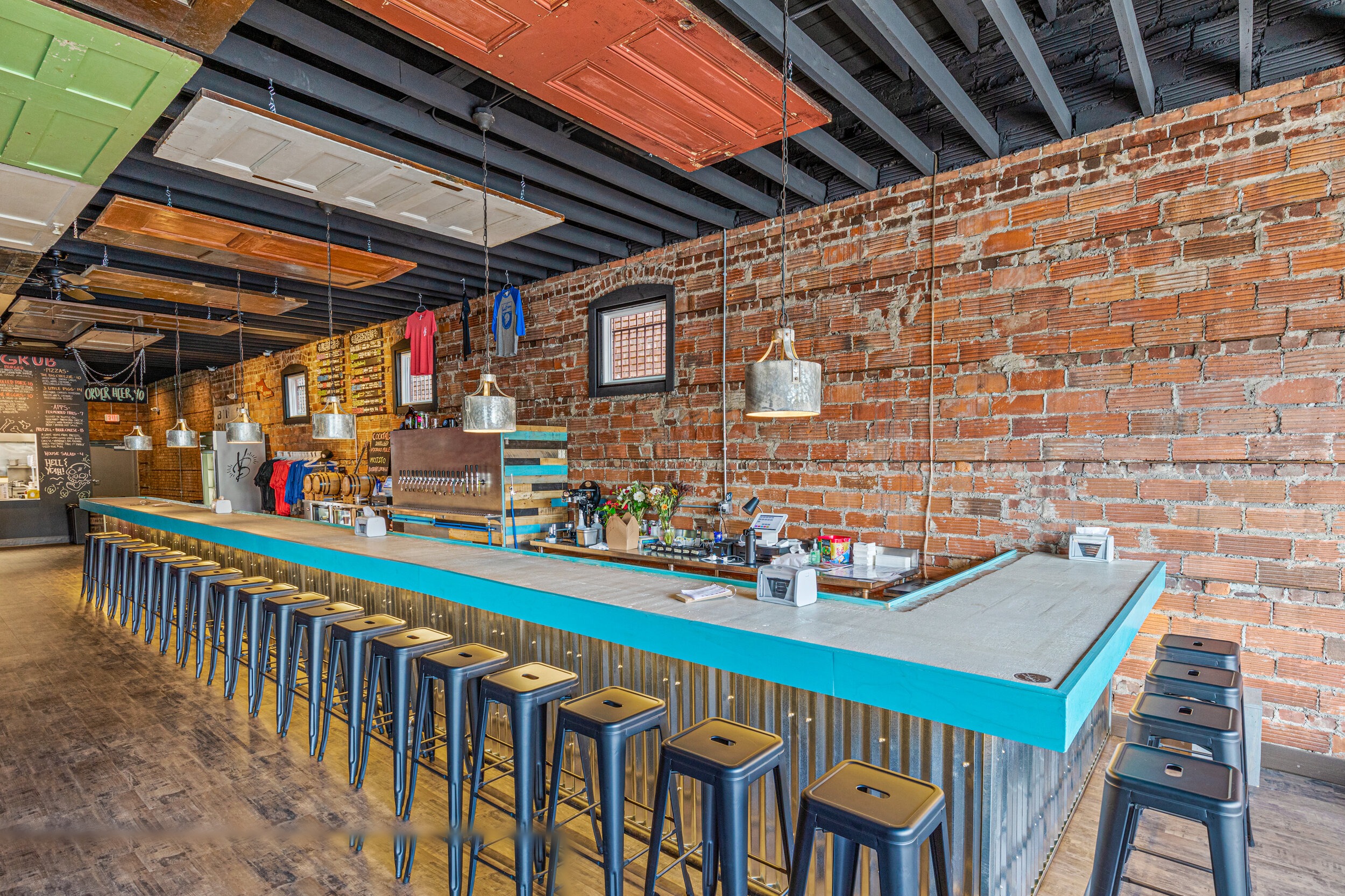 Teal-painted bar with concrete countertop, corrugated metal base, metal stools, and exposed brick wall — VooDoo Brewery by Skoda Construction