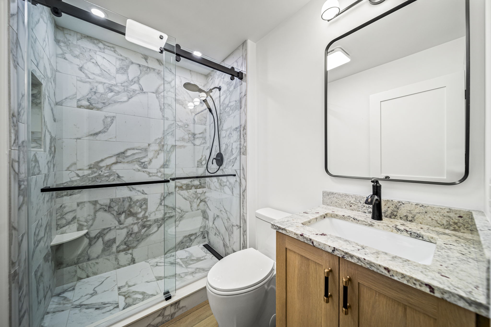 Bathroom with dramatic marble-look porcelain shower, barn-door glass enclosure, and natural oak vanity — custom home by Skoda Construction, Northeast Ohio