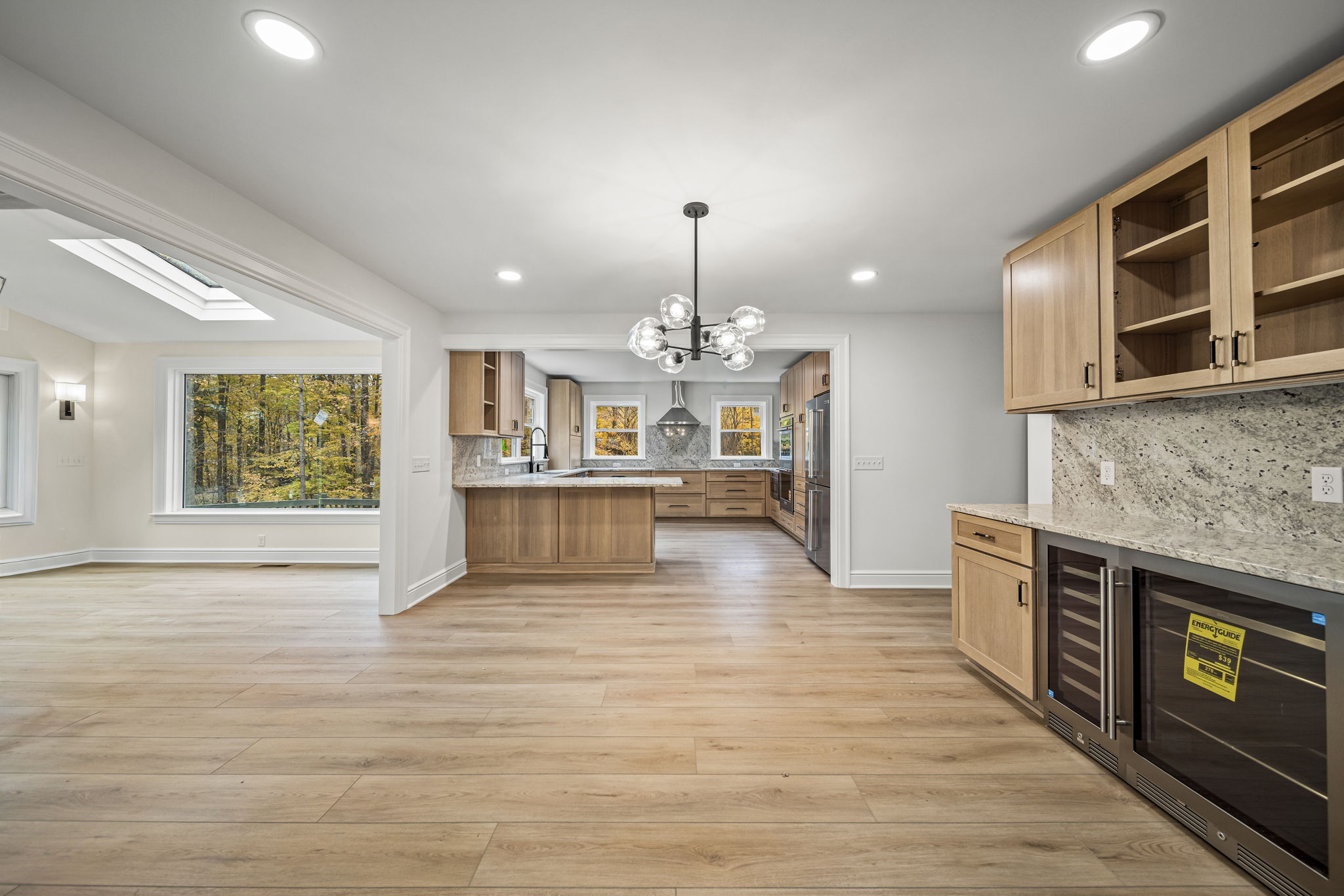 Open-concept great room with sputnik chandelier, wine bar, and natural oak kitchen overlooking autumn woods — Skoda Construction, Northeast Ohio