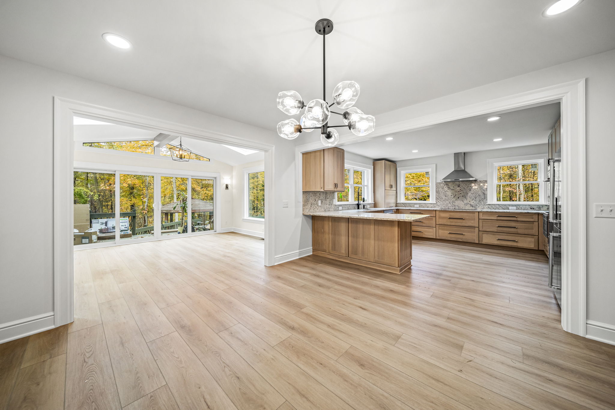 Spectacular wide-angle open floor plan showing dining area with sputnik chandelier, natural oak kitchen, and four-season sunroom with autumn woodland views — custom home by Skoda Construction, Northeast Ohio