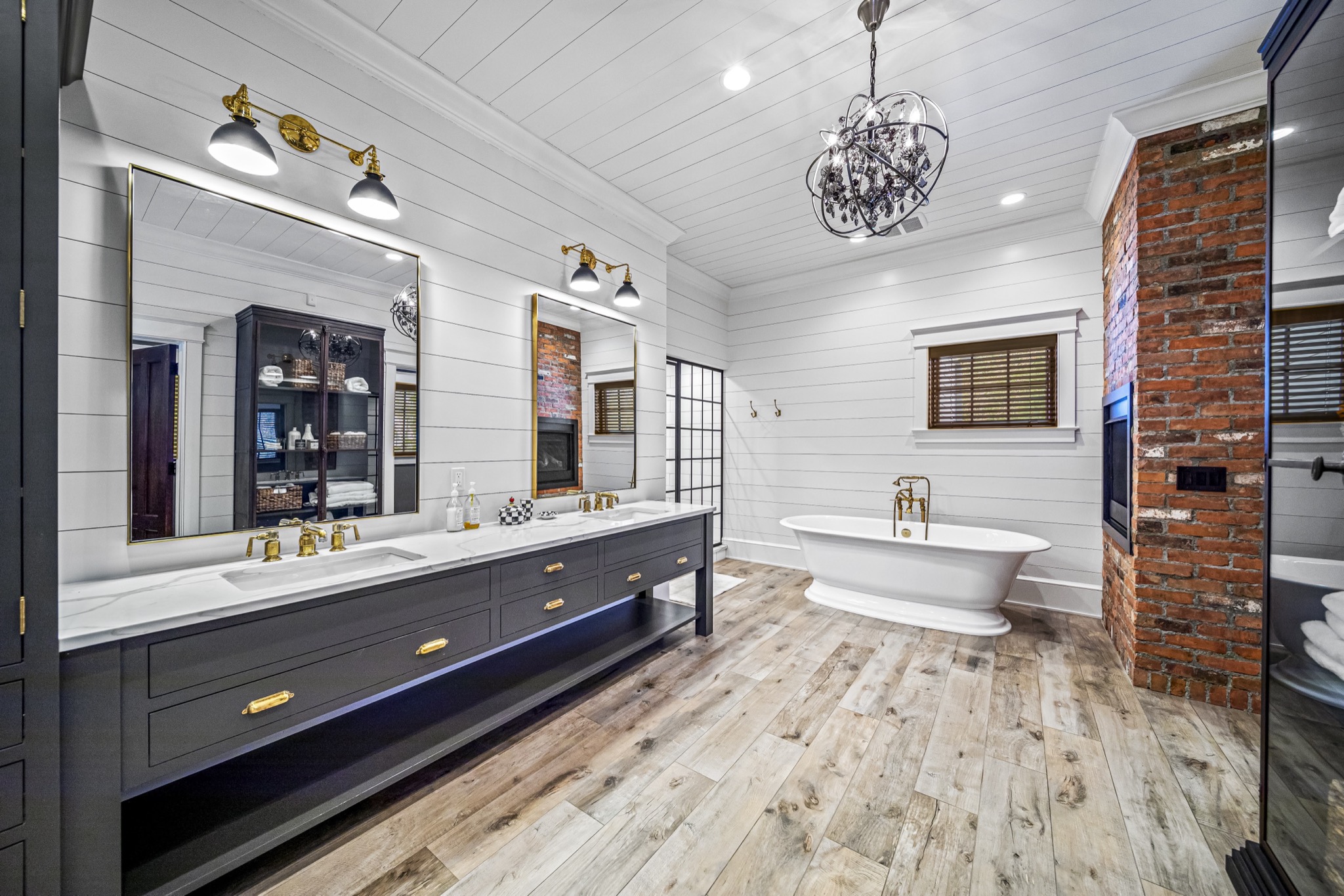 Luxury master bathroom with freestanding soaking tub, crystal chandelier, LED-lit double vanity, exposed brick column, and gold fixtures — custom home by Skoda Construction, Northeast Ohio