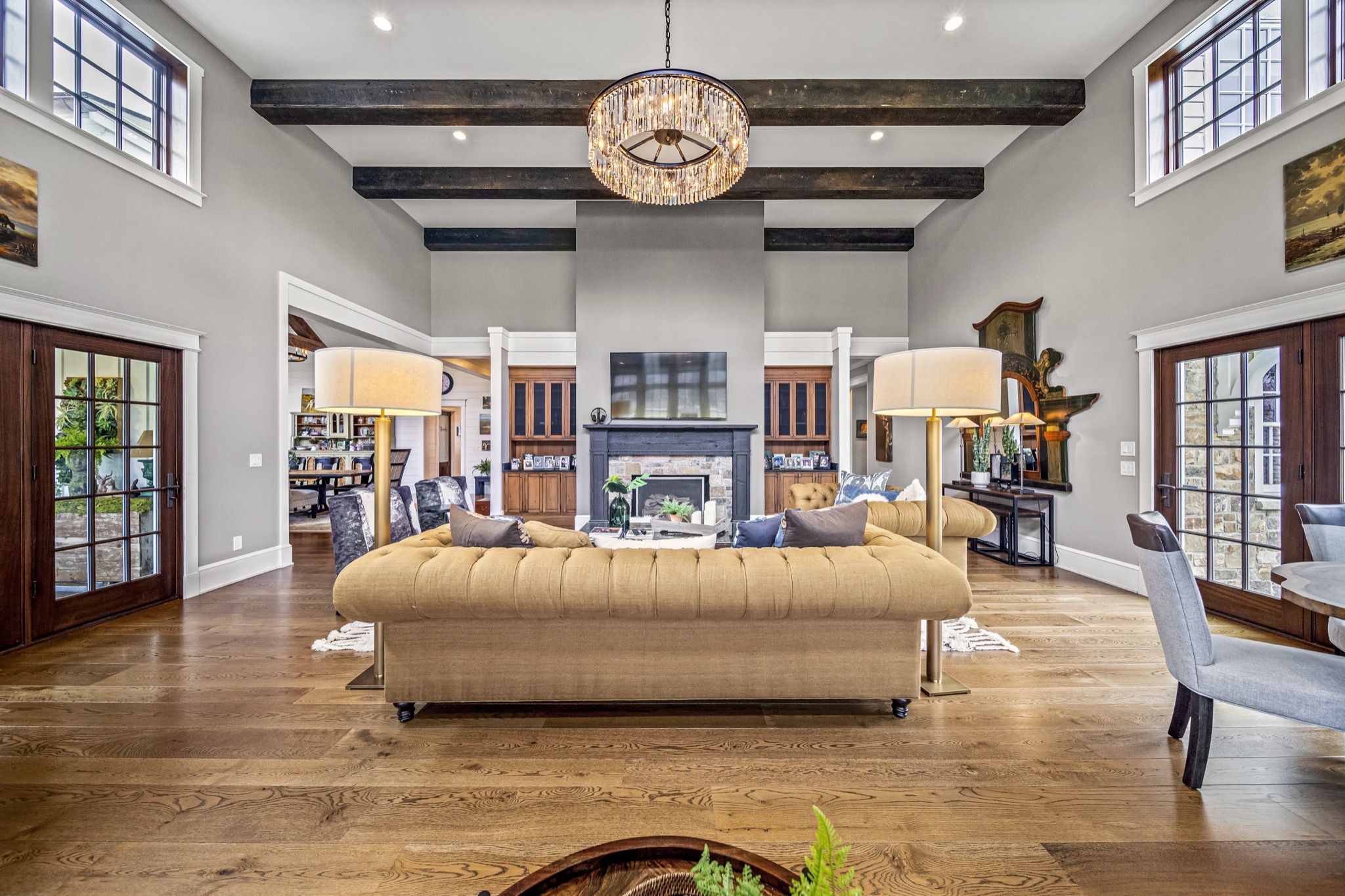Symmetrical living room with tufted chesterfield sofa, crystal drum chandelier, dark ceiling beams, and flanking French doors — luxury home by Skoda Construction, Northeast Ohio