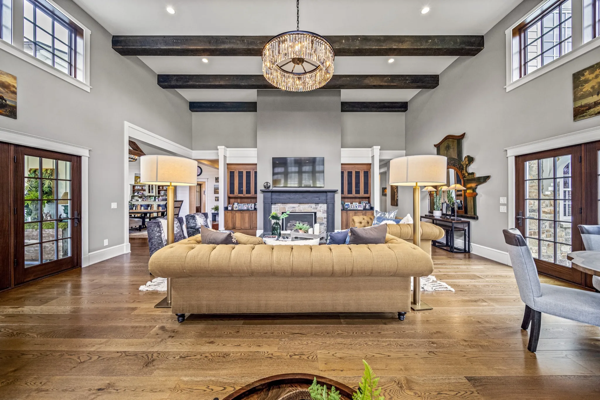 Symmetrical living room with tufted chesterfield sofa, crystal drum chandelier, dark ceiling beams, and flanking French doors — luxury home by Skoda Construction, Northeast Ohio