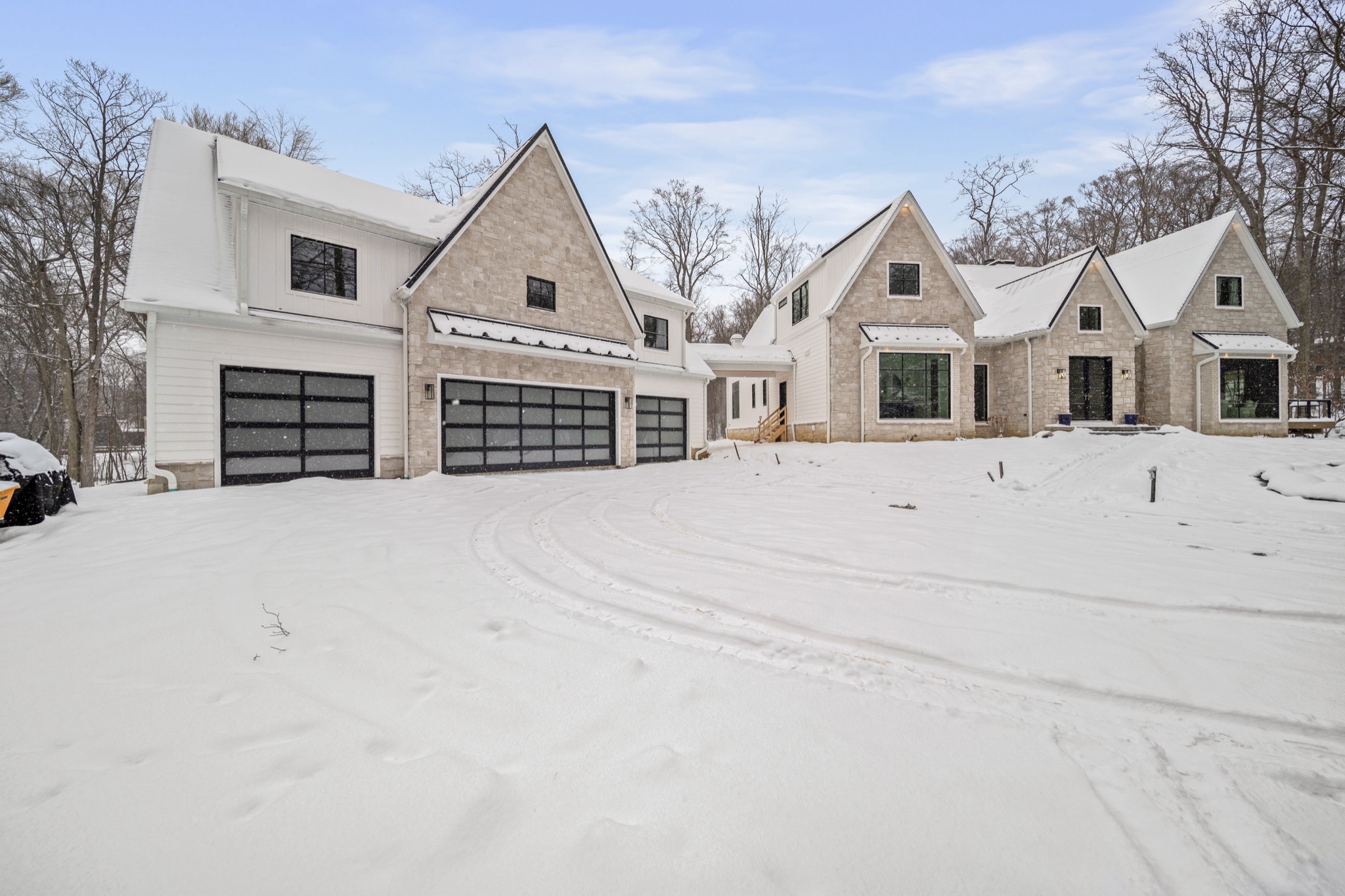 Modern luxury estate from the garage approach showing triple glass garage doors, natural stone gables, and snow-covered wooded lot — custom home by Skoda Construction, Northeast Ohio