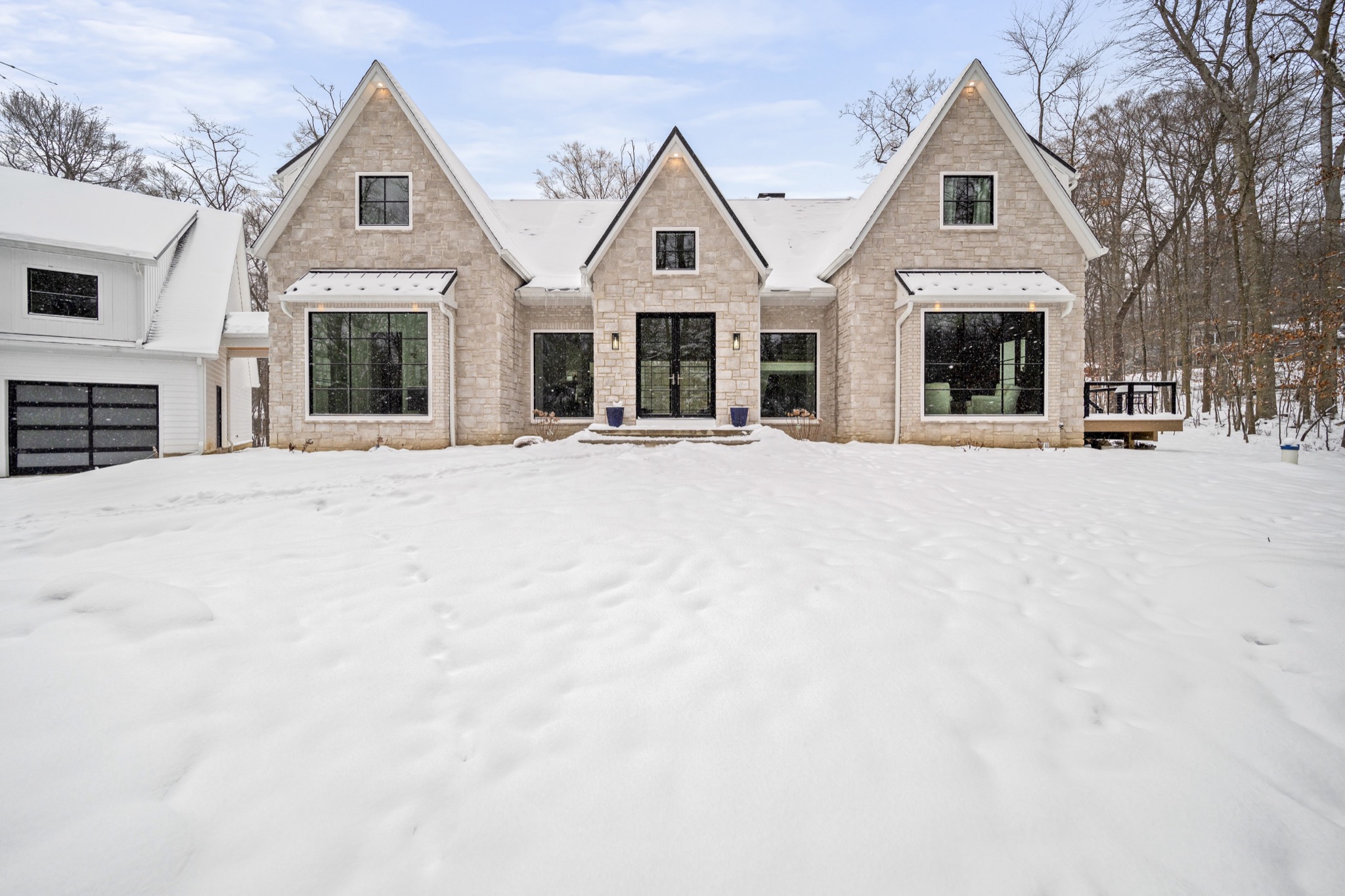Symmetrical stone-clad modern farmhouse facade with twin peaked gables and black-framed windows surrounded by fresh snowfall — custom home by Skoda Construction