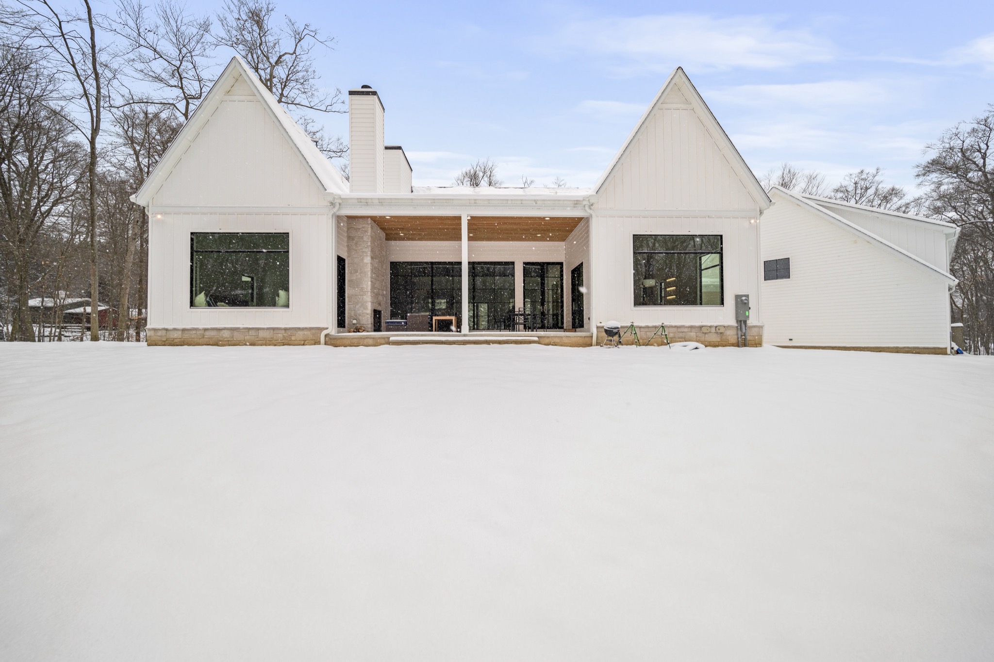 Rear elevation of modern luxury estate featuring covered porch, natural stone chimney, and walls of glass overlooking a private wooded lot blanketed in snow — Skoda Construction