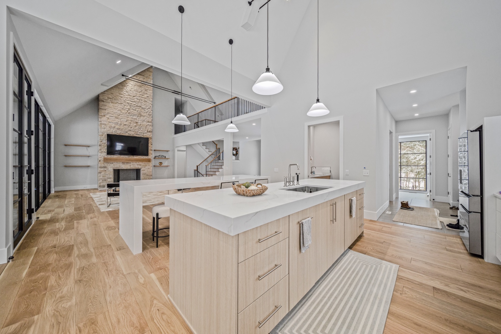 Gourmet kitchen island with white quartz countertop beneath vaulted ceilings, with natural stone fireplace wall and open loft walkway visible beyond — Skoda Construction luxury home