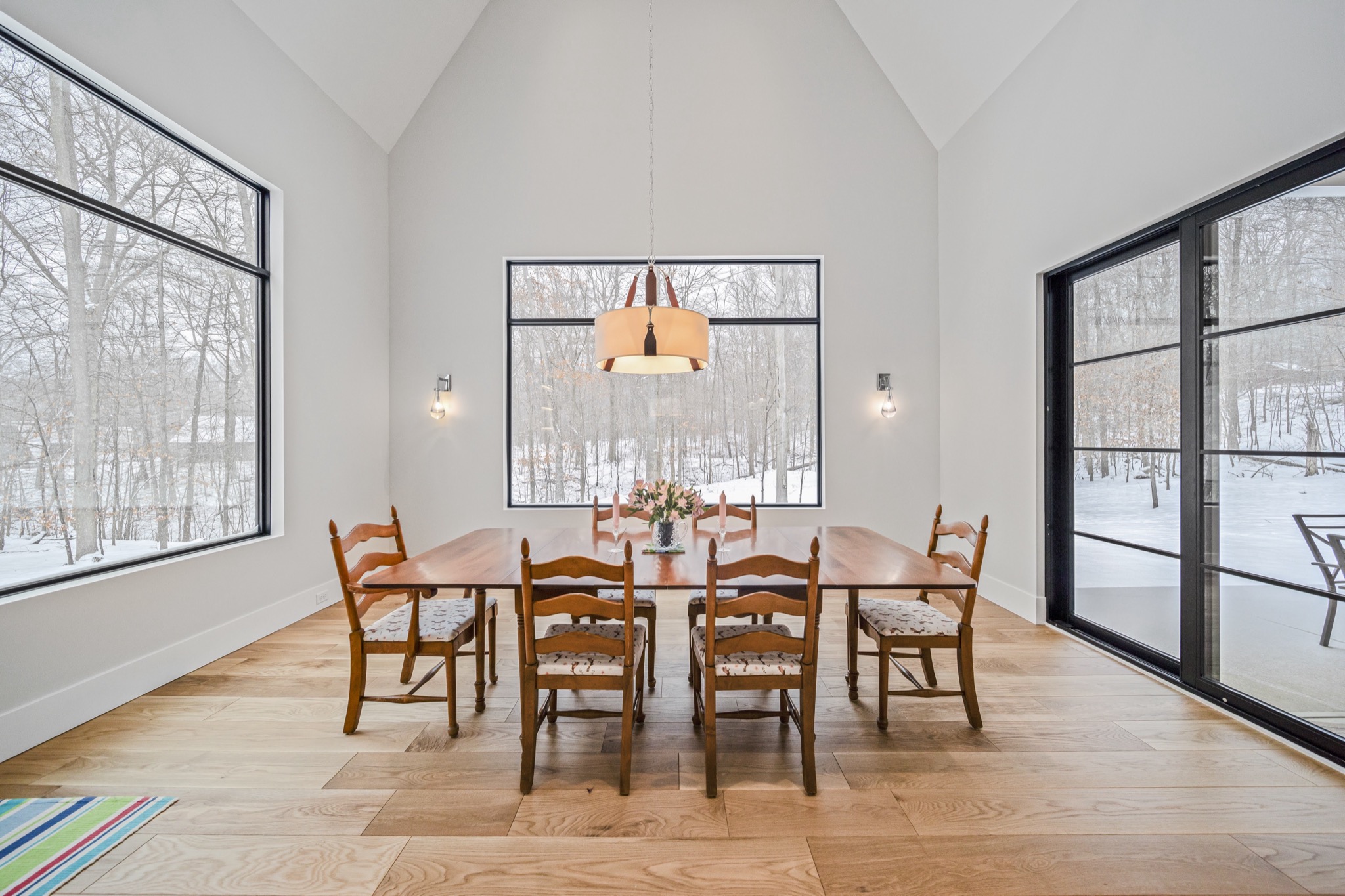 Light-filled formal dining room with floor-to-ceiling windows overlooking snowy woodland, vaulted ceiling and modern pendant chandelier — Skoda Construction custom estate