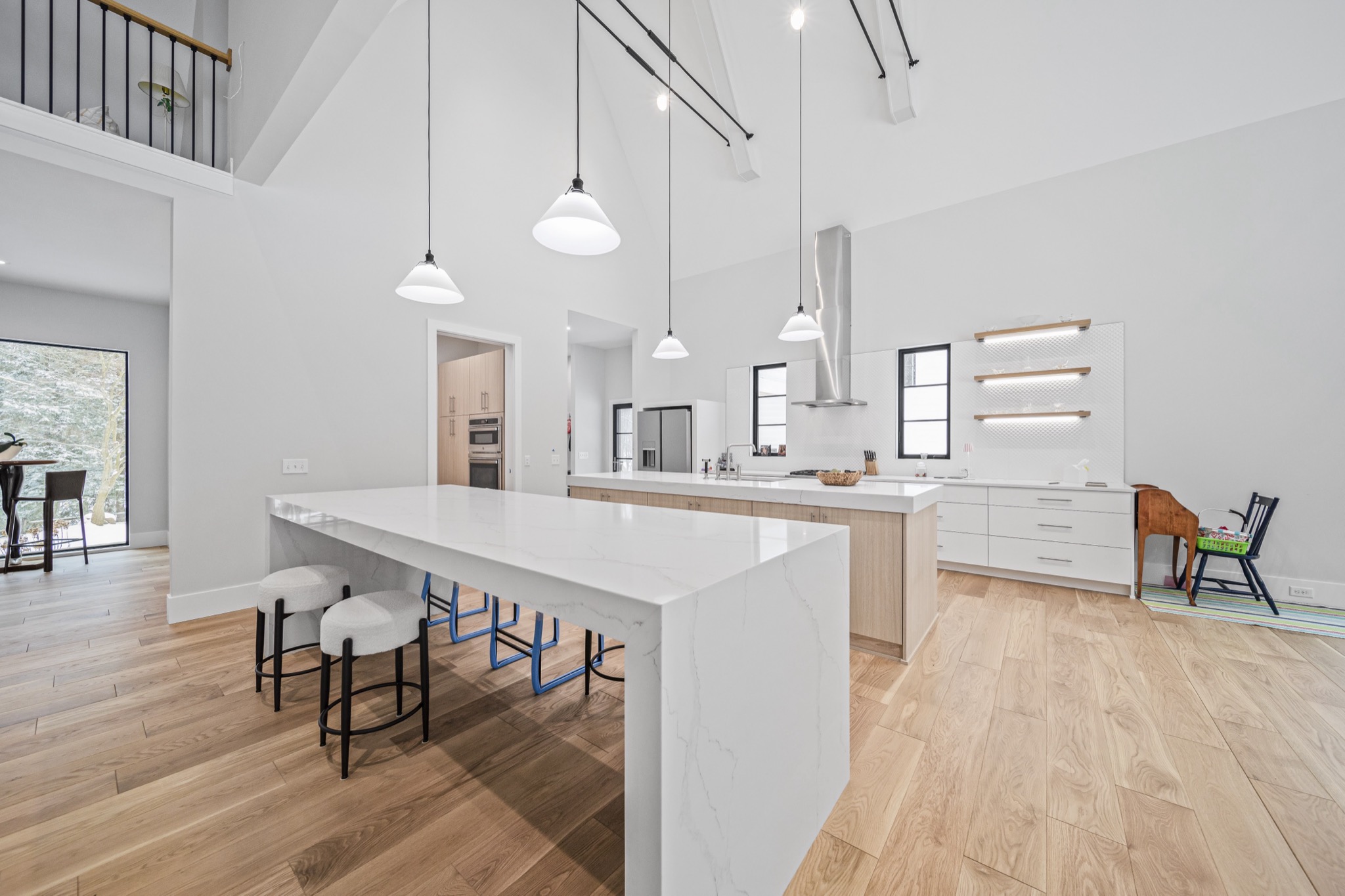 Waterfall-edge quartz kitchen island with modern bar seating beneath towering vaulted ceiling with pendant lighting and floating wood shelves — Skoda Construction luxury home