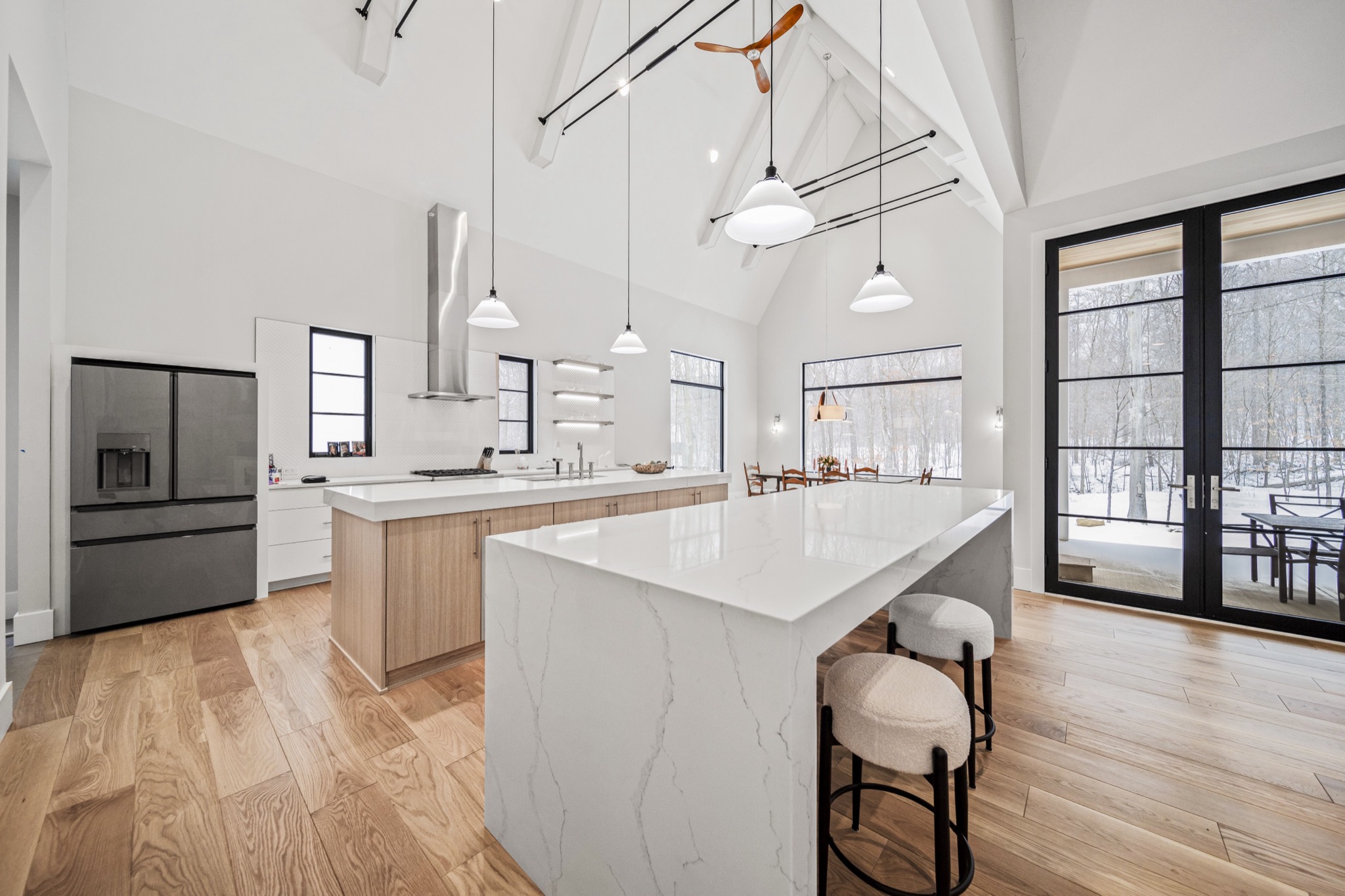 Expansive modern kitchen with dual islands, waterfall marble countertops, floor-to-ceiling glass walls, and cathedral ceiling with exposed structural beams — Skoda Construction custom estate, Northeast Ohio