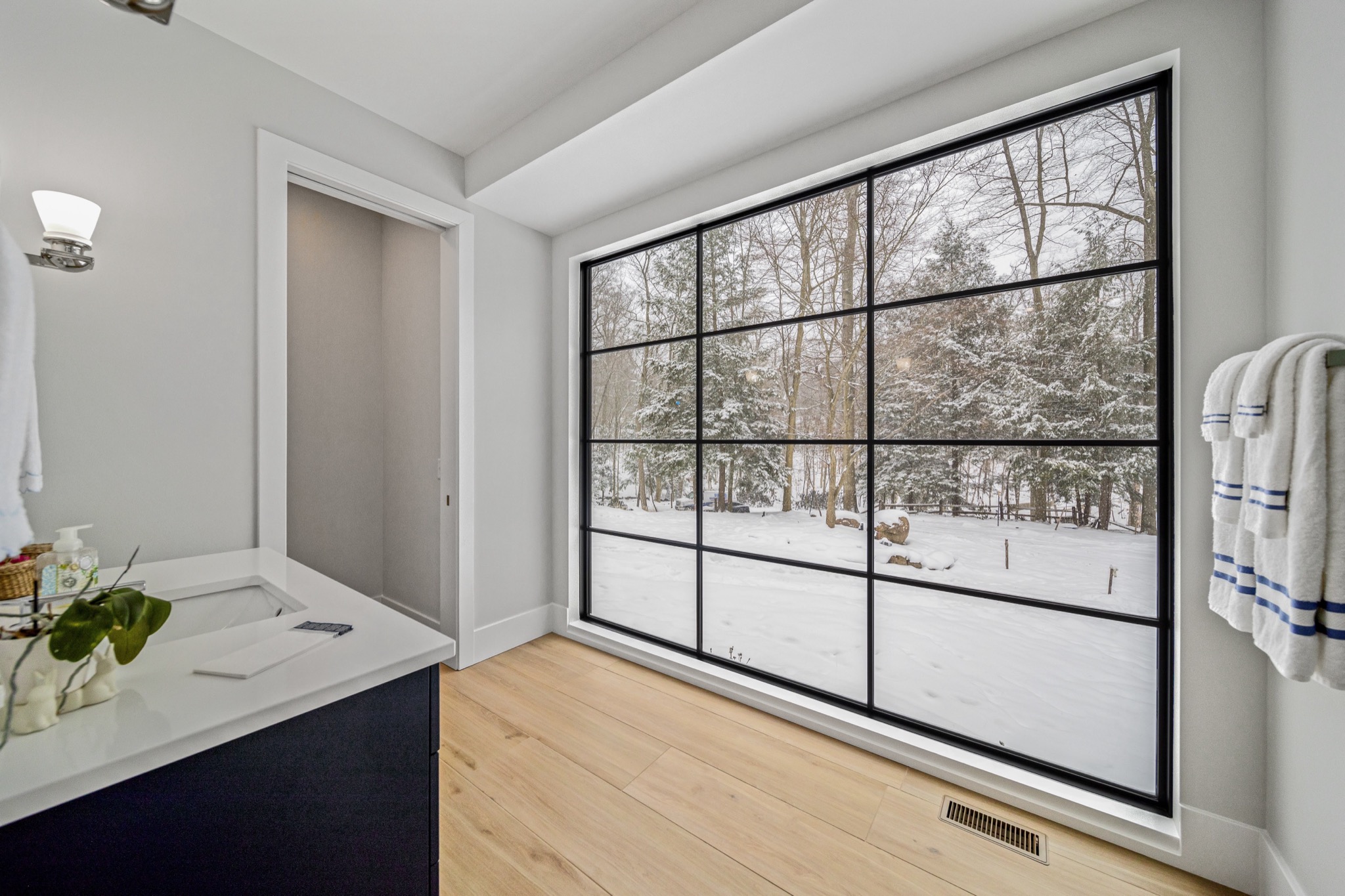 Bathroom with floor-to-ceiling grid-paned window overlooking snow-covered woodland, dark vanity, and quartz countertop — modern luxury home by Skoda Construction, Northeast Ohio