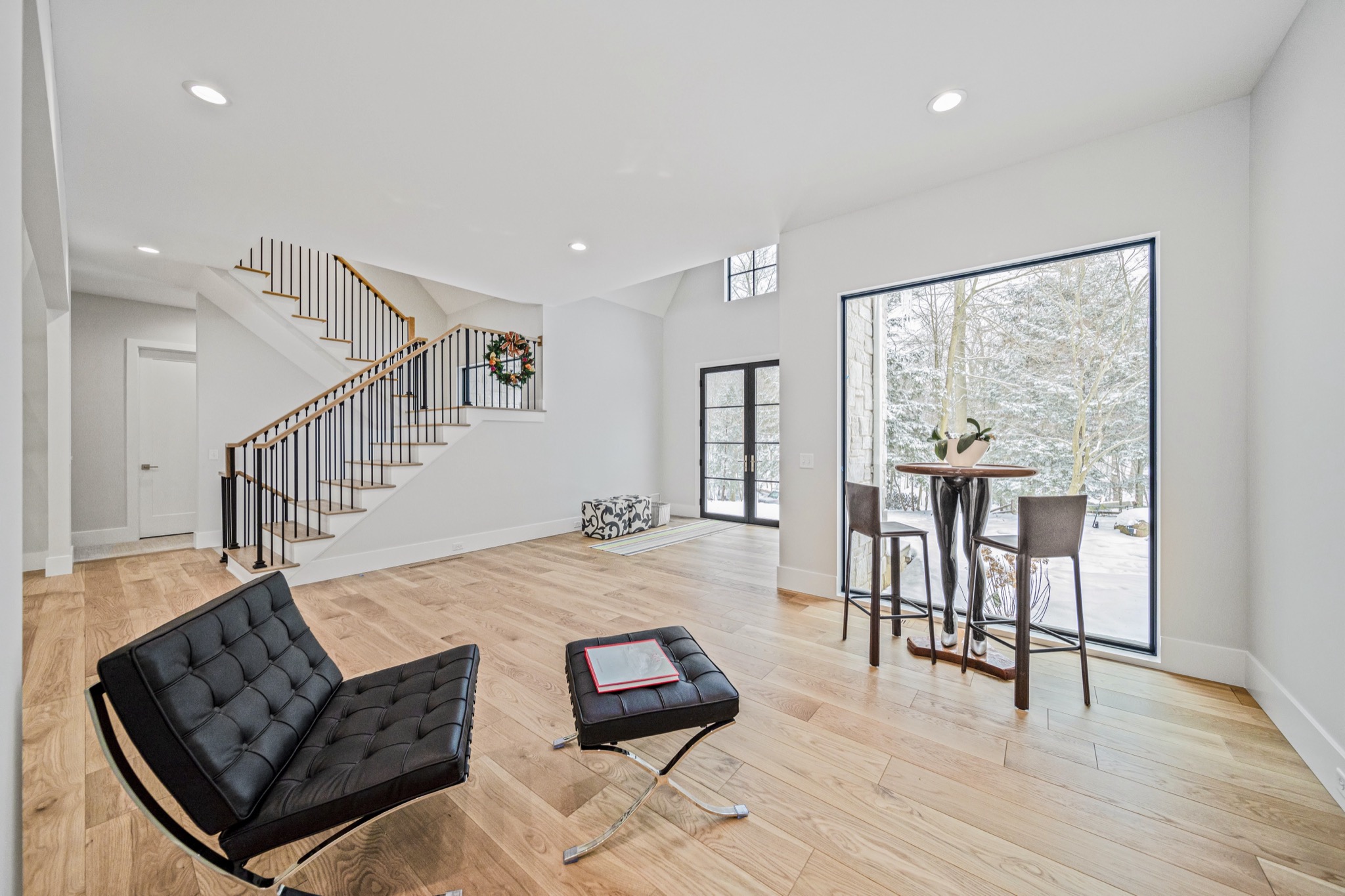 Lower level lounge with Barcelona chair, modern staircase, floor-to-ceiling glass doors to covered patio, and snowy woodland views — luxury estate by Skoda Construction, Northeast Ohio