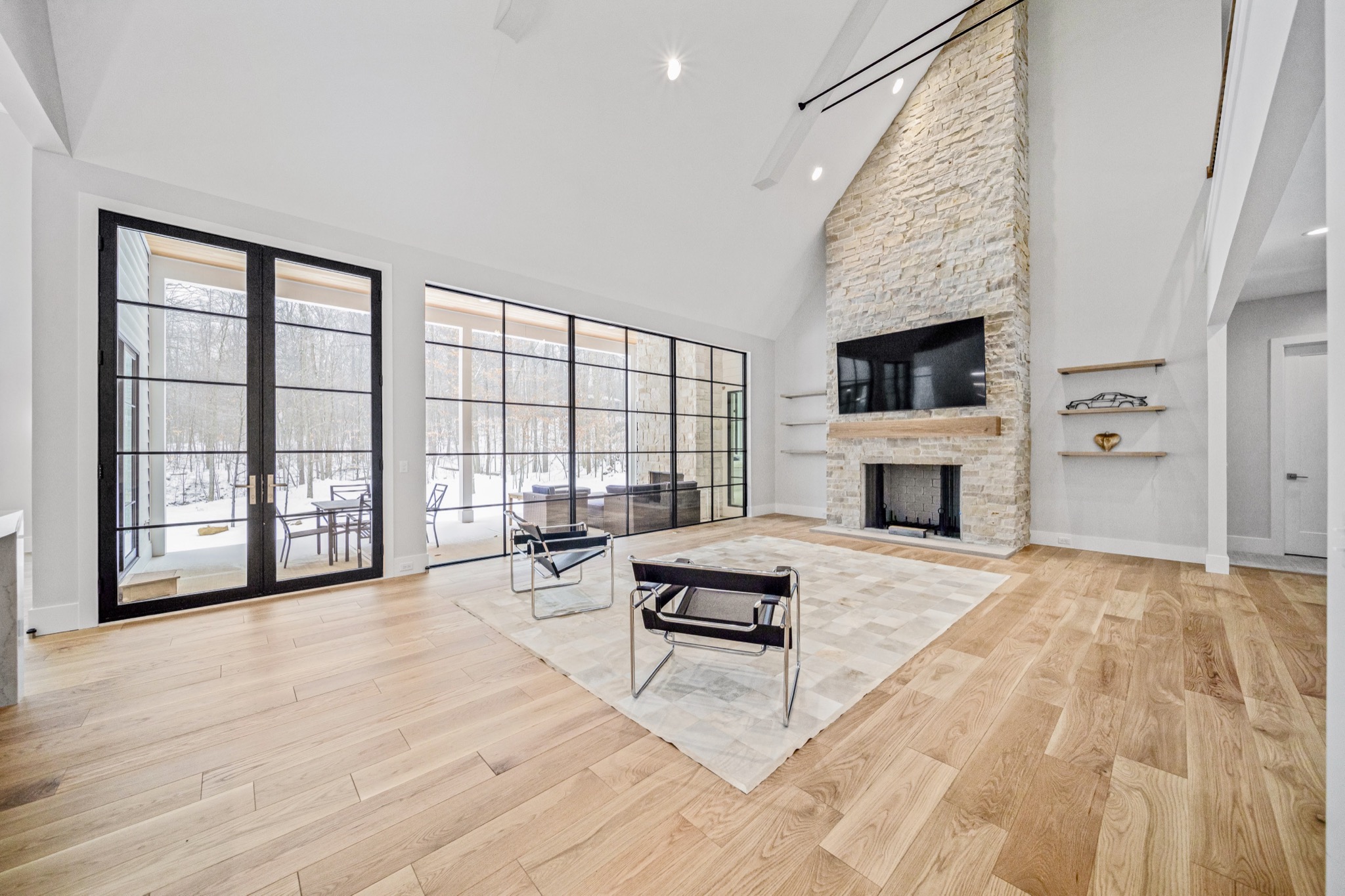 Modern great room with floor-to-ceiling glass wall revealing snow-covered woodland, natural stone fireplace, and covered outdoor living space beyond — Skoda Construction luxury estate