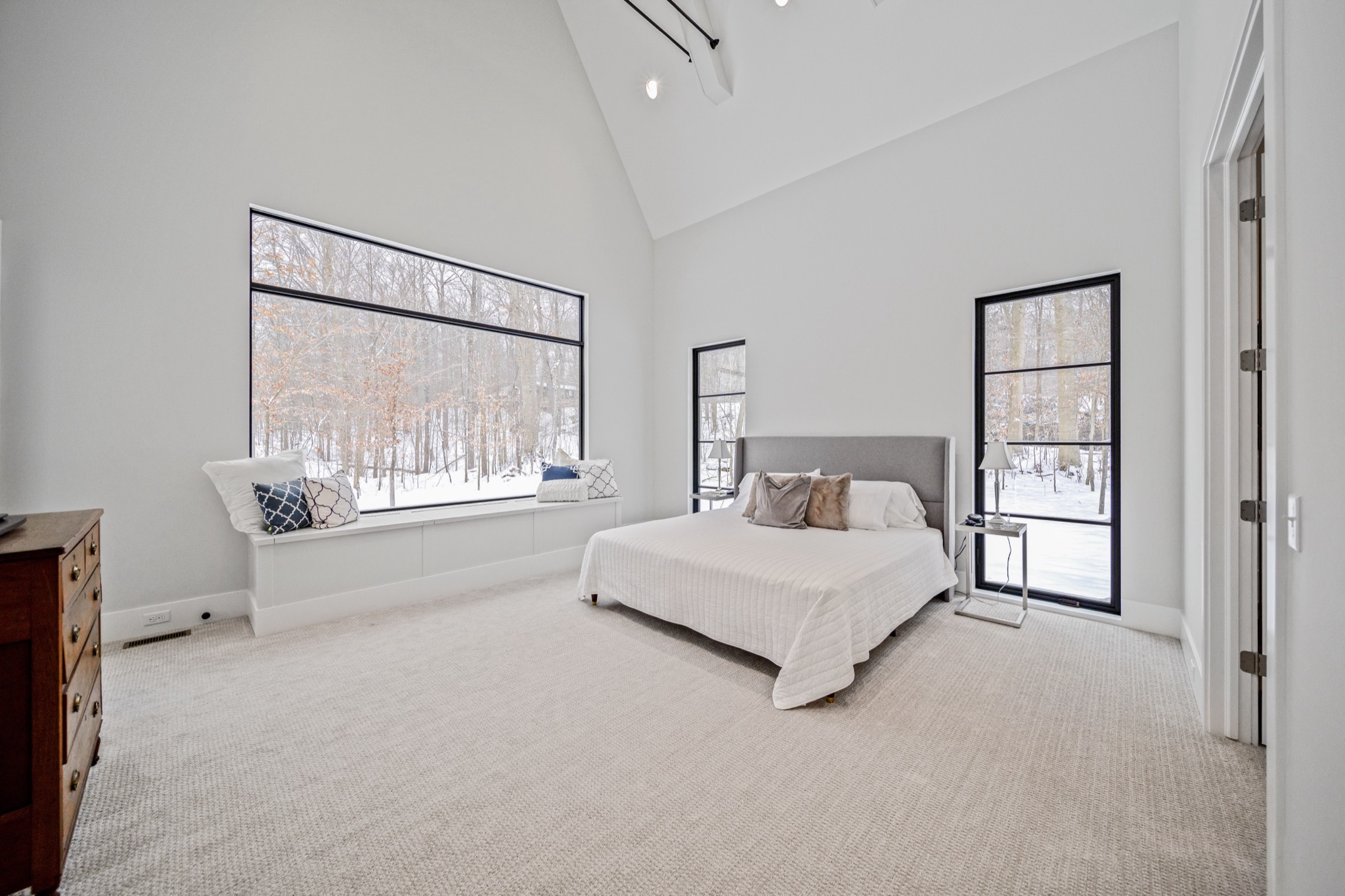 Luxurious primary bedroom with vaulted ceiling, oversized picture window with built-in window seat, and floor-to-ceiling glass door overlooking a private wooded setting — Skoda Construction custom home