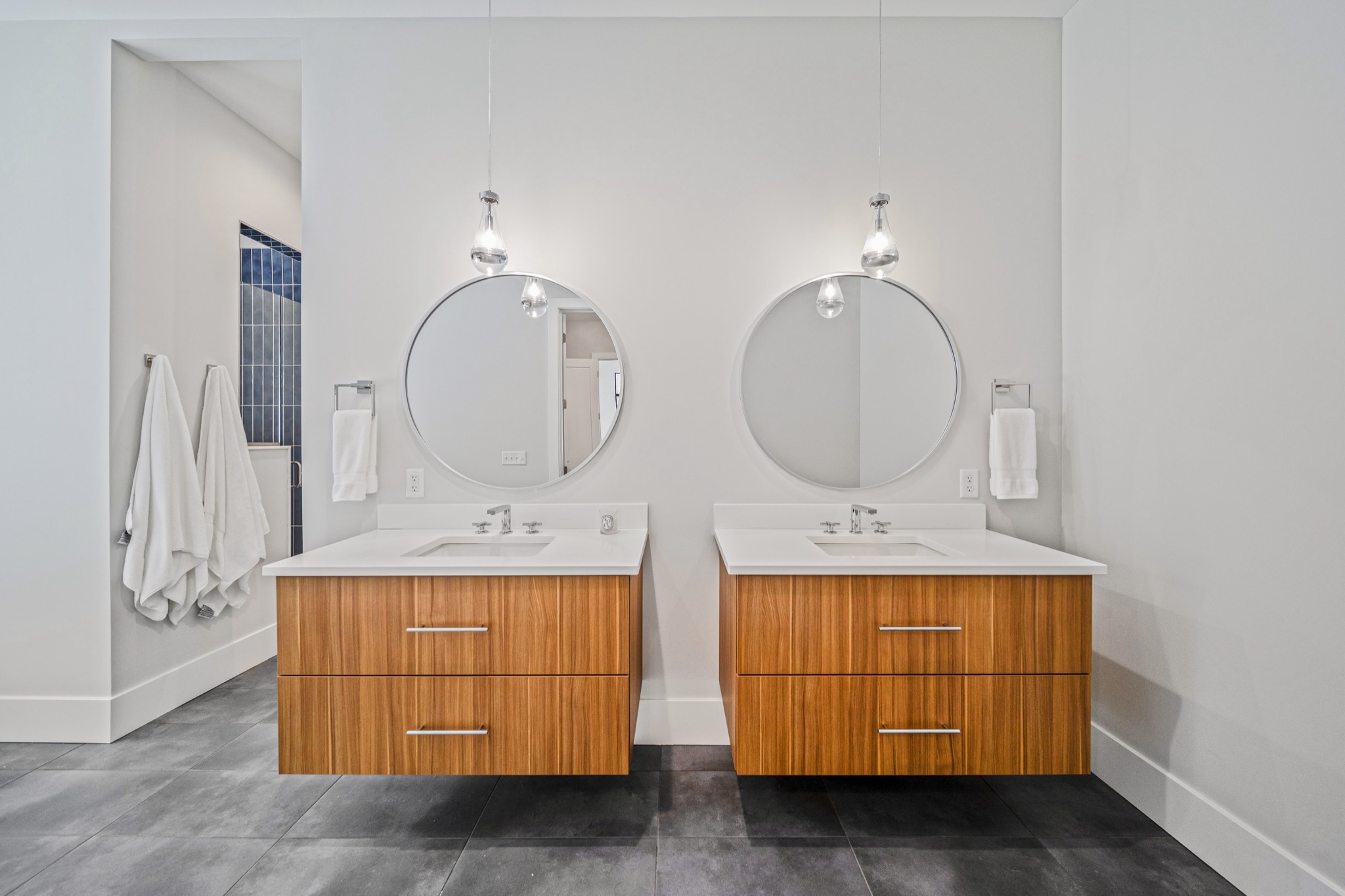 Modern primary bathroom with dual floating walnut vanities, oversized round mirrors, and globe pendant lighting on dark slate-look tile floors — Skoda Construction luxury home