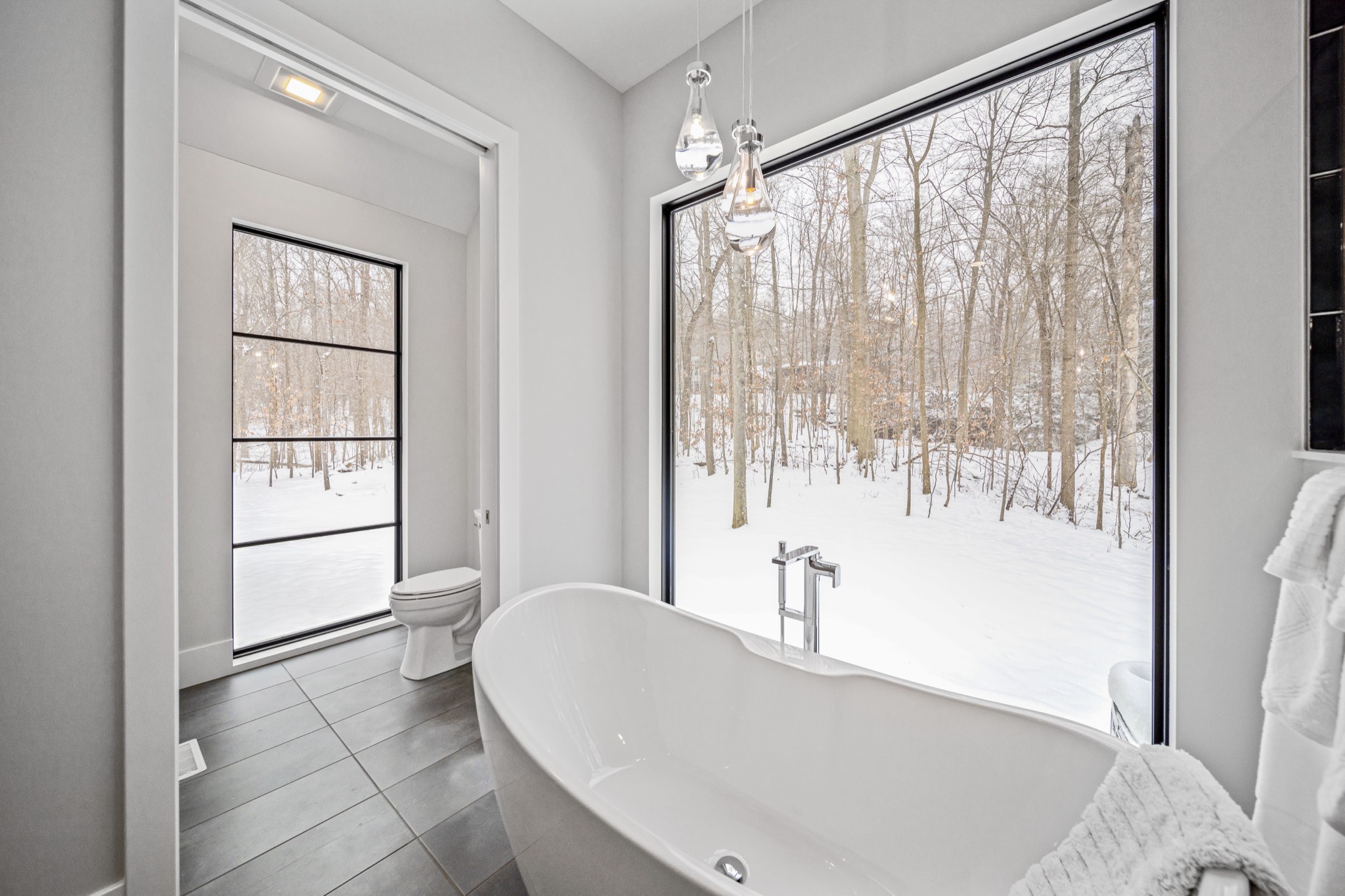 Freestanding soaking tub beneath an expansive picture window with unobstructed views of a snow-covered private woodland — luxury bathroom by Skoda Construction