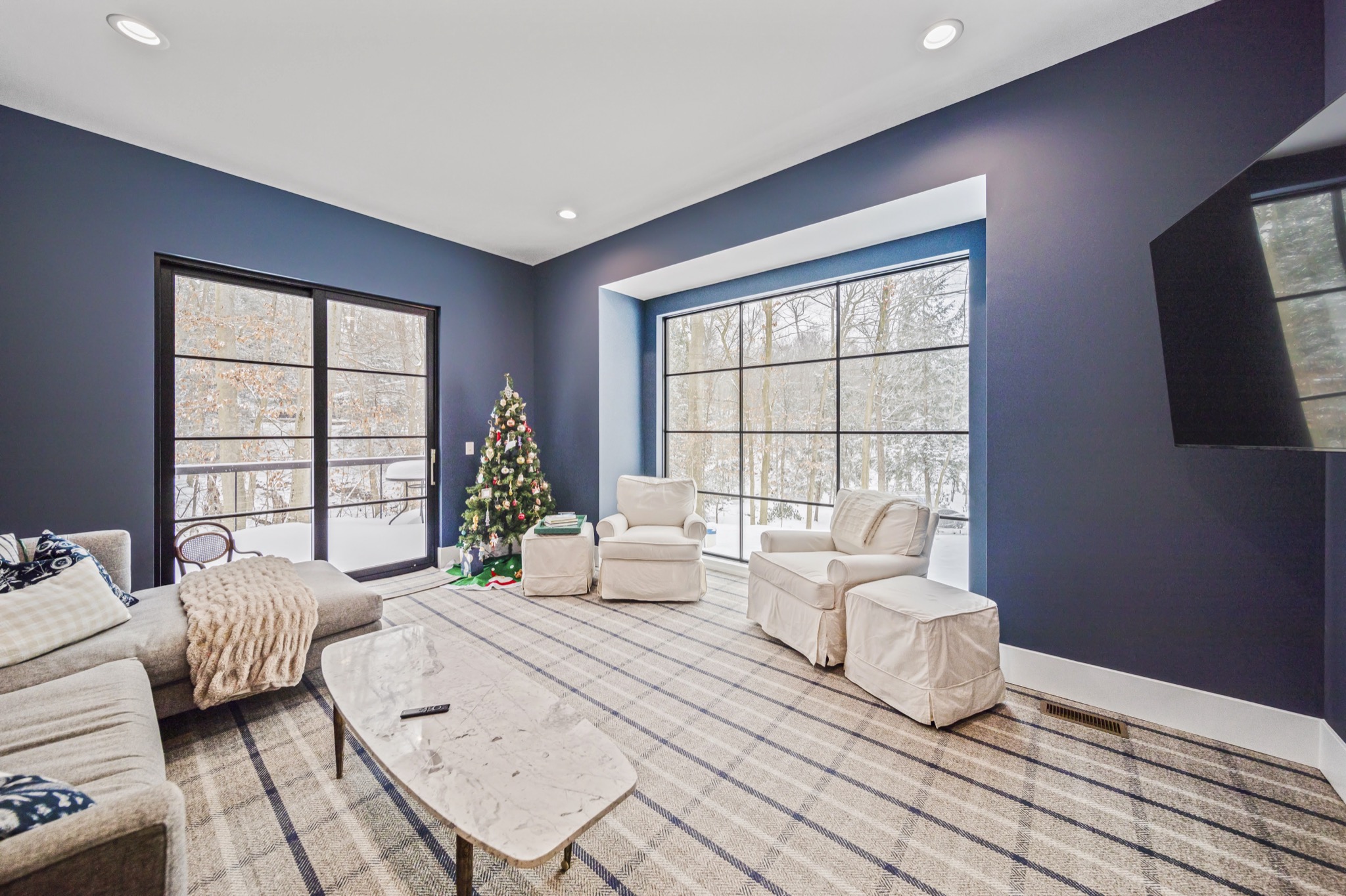 Second-floor media room with bold navy walls, floor-to-ceiling grid windows overlooking snowy woodland, and cream seating on plaid rug — luxury estate by Skoda Construction, Northeast Ohio