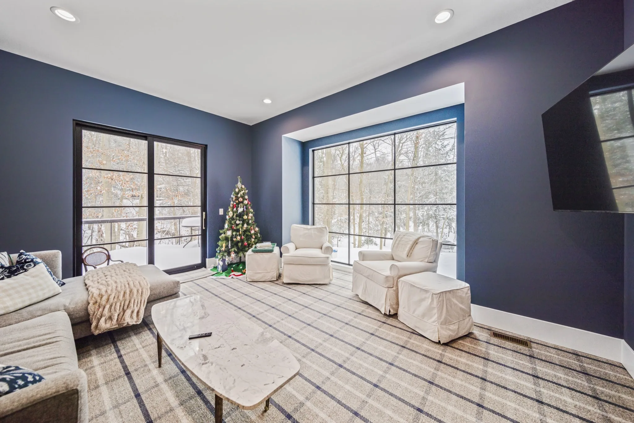 Second-floor media room with bold navy walls, floor-to-ceiling grid windows overlooking snowy woodland, and cream seating on plaid rug — luxury estate by Skoda Construction, Northeast Ohio