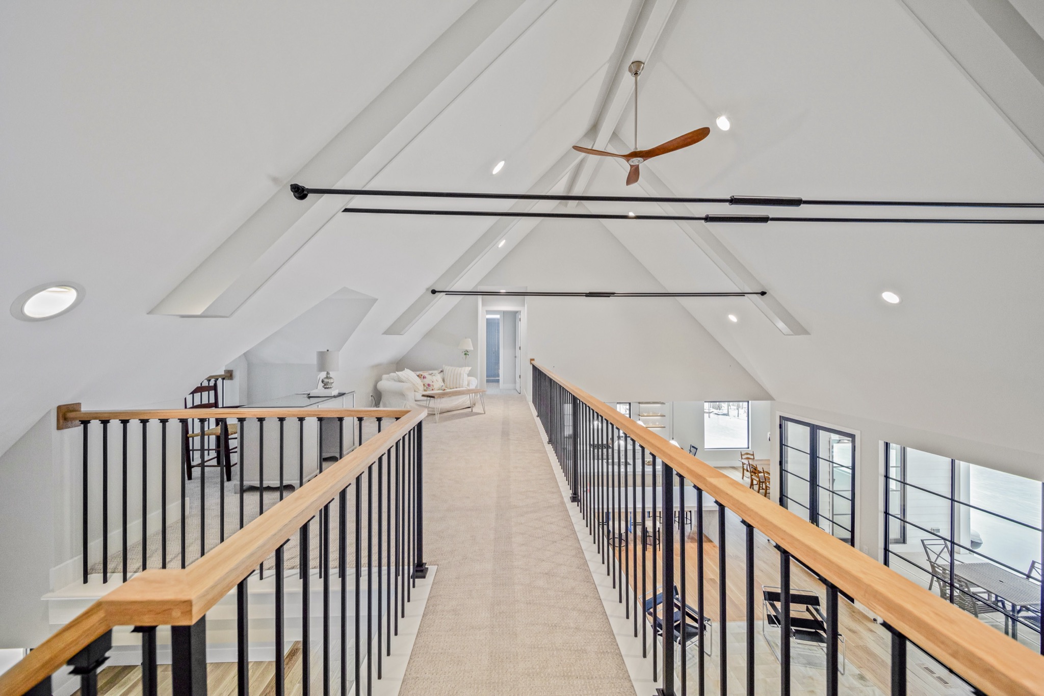 Open loft walkway with iron-and-wood railing beneath a soaring cathedral ceiling with exposed structural beams and modern ceiling fan — Skoda Construction custom estate