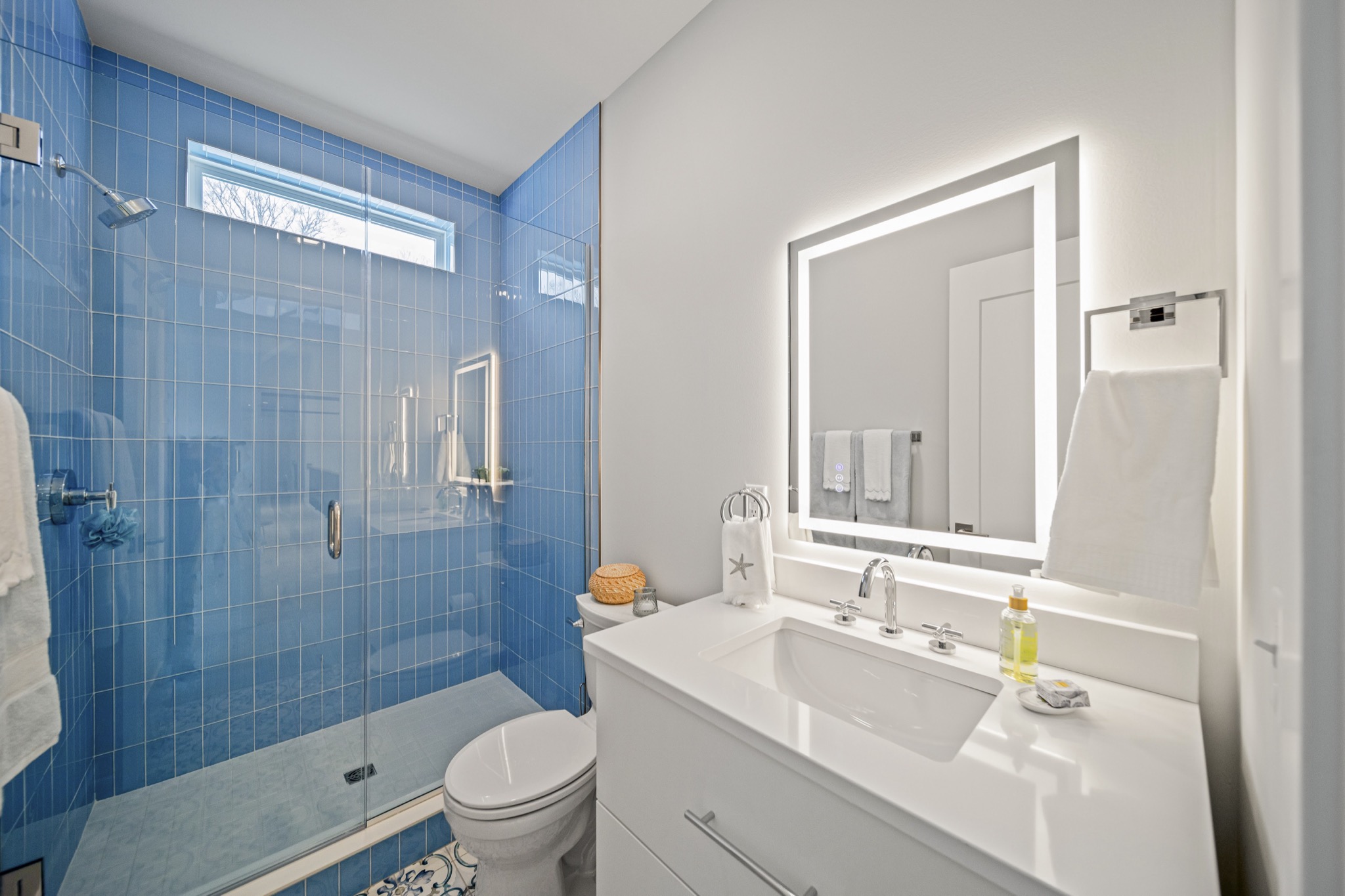 Contemporary bathroom with vibrant blue tile walk-in shower, LED-backlit vanity mirror, and mosaic tile flooring — luxury custom home by Skoda Construction, Northeast Ohio