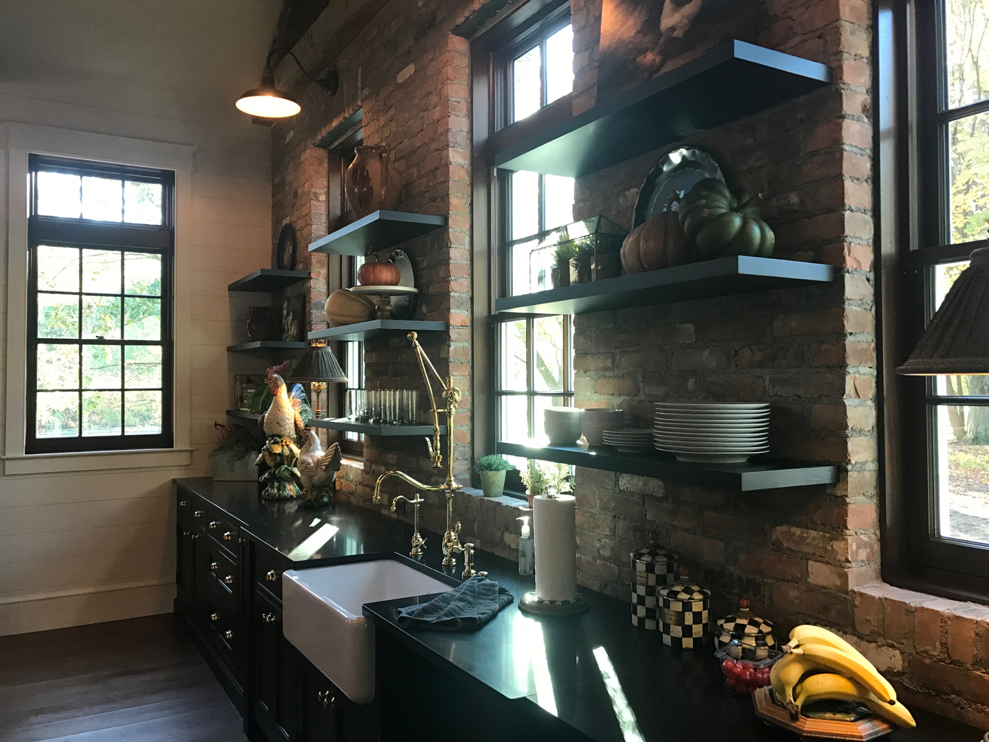 Farmhouse kitchen with exposed brick wall, dark cabinetry, open wood shelving, brass bridge faucet, and apron-front sink — custom home renovation by Skoda Construction, Northeast Ohio